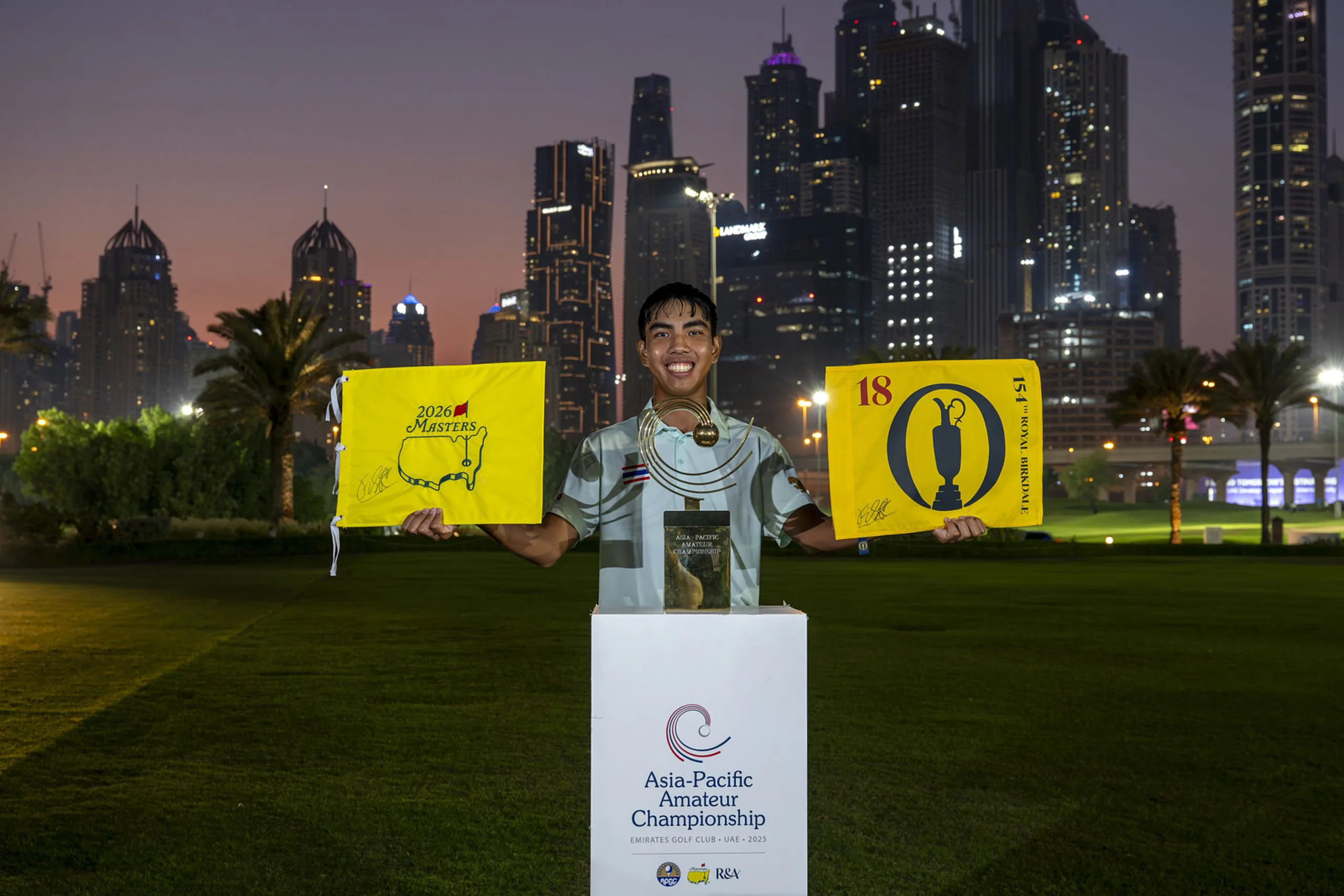 Fifa Laopakdee of Thailand, winner of the 2025 Asia-Pacific Amateur Championship, lifts The 154th Open Championship flag and the Masters flag alongside the Asia-Pacific Amateur Championship trophy in front of the Dubai Marina skyline after winning the 2025 Asia-Pacific Amateur Championship following a Play-Off at the 2025 Asia-Pacific Amateur Championship being played at the Emirates Golf Club Majlis Course in Dubai, United Arab Emirates on Sunday 26 October 2025. Photograph by AAC.